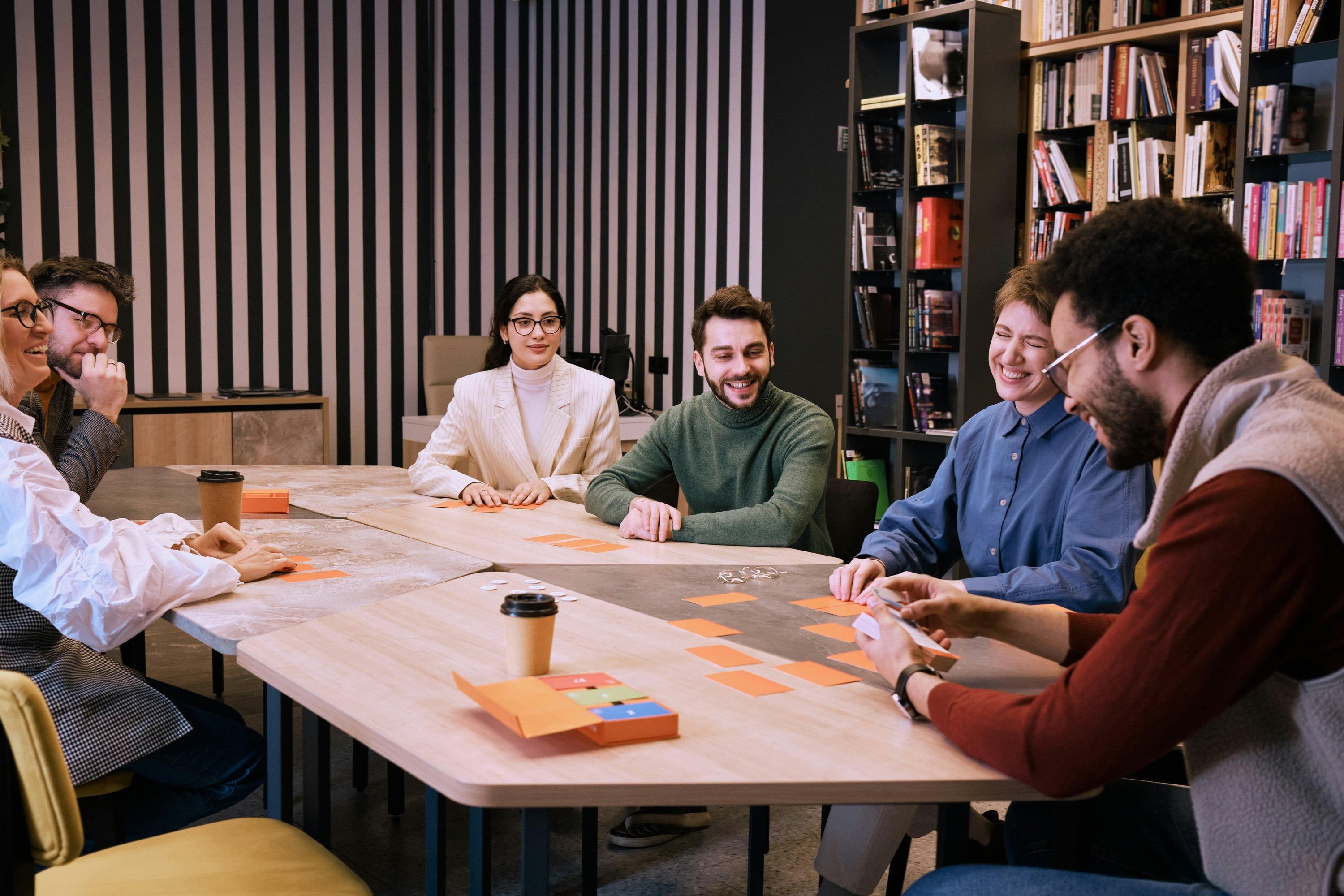 Équipe diverse collaborant lors d'une réunion autour d'une table en bois, travaillant avec des notes adhésives orange dans un espace moderne avec mur rayé et bibliothèque