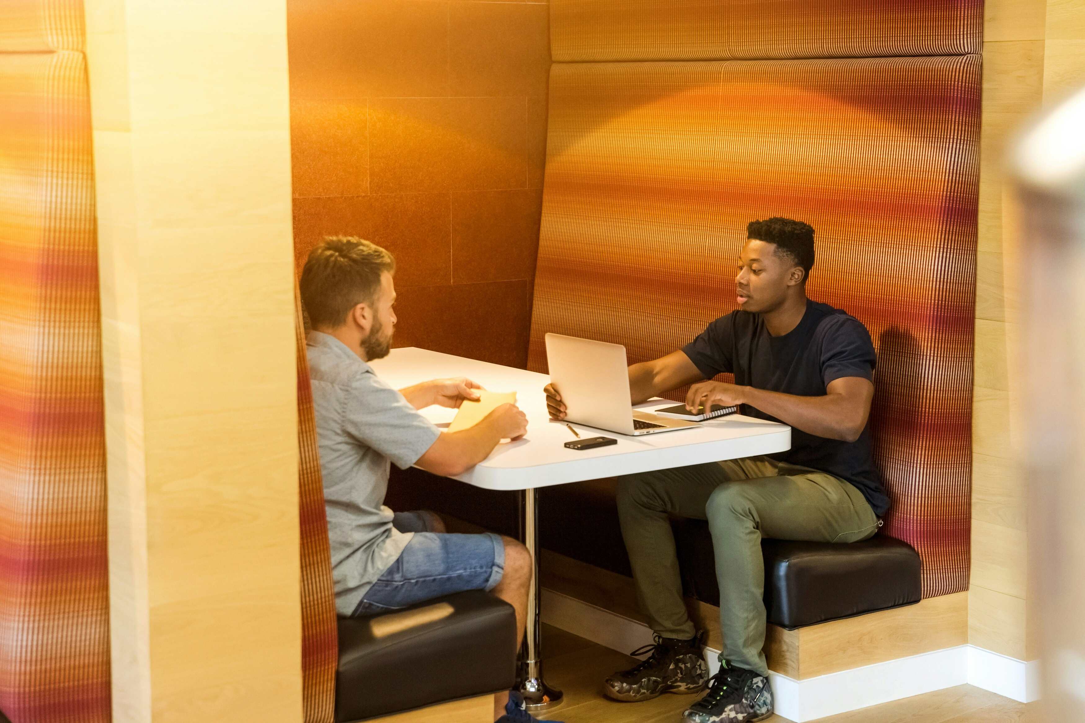 Deux hommes discutant dans un espace de travail moderne avec des murs orangés, l'un utilisant un ordinateur portable sur une table blanche, illustrant la collaboration et les solutions d'accès data