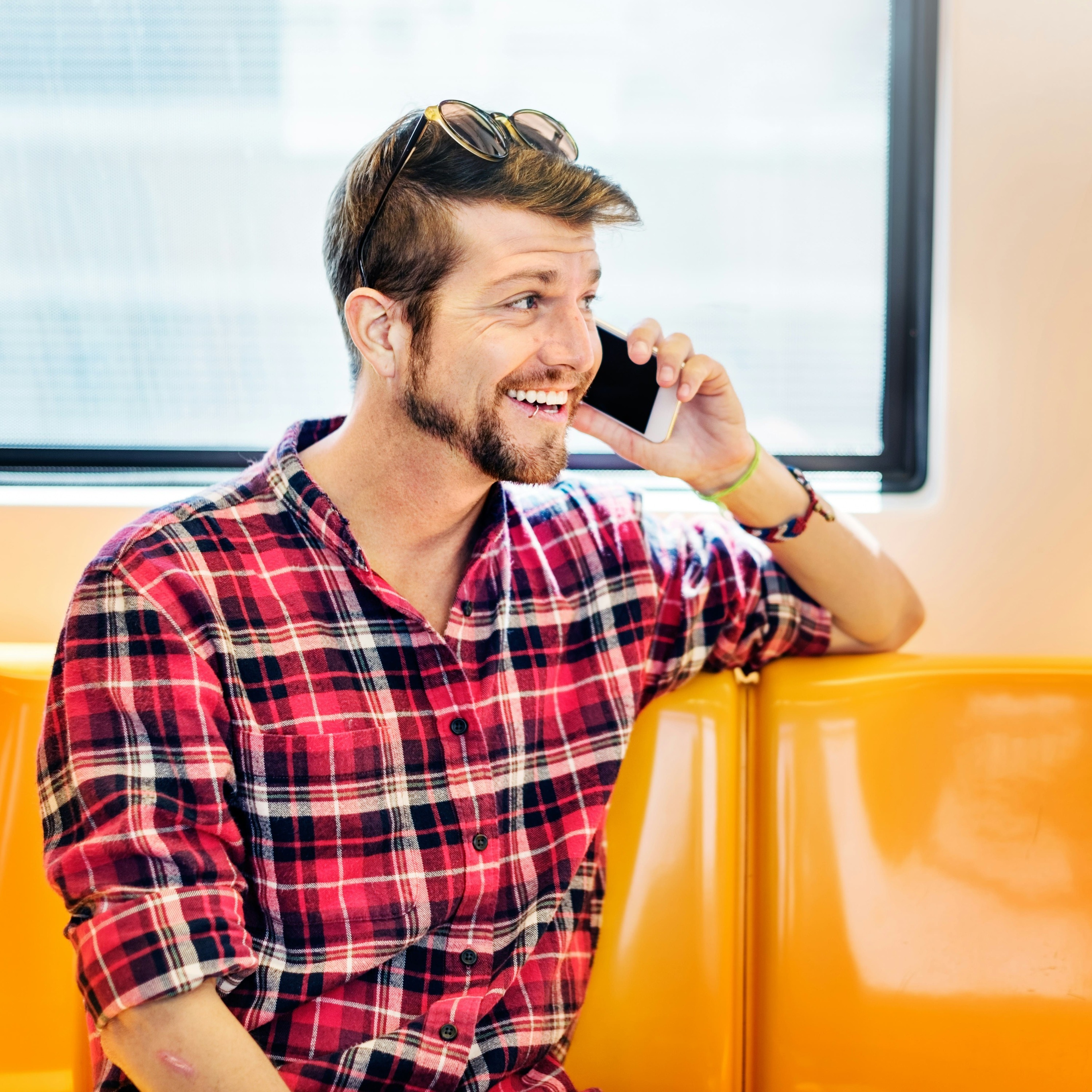 Jeune homme souriant avec une chemise à carreaux rouges parlant au téléphone, assis sur un siège orange, illustrant la communication mobile moderne